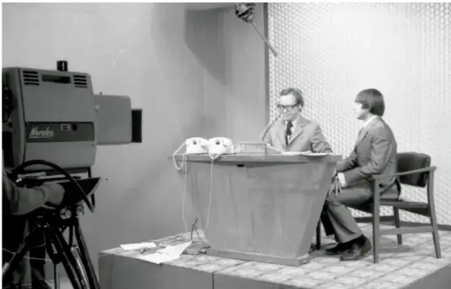 Fotografía en blanco y negro de un set de televisión con un camarógrafo, un conductor y una conductora sentados en un escritorio.