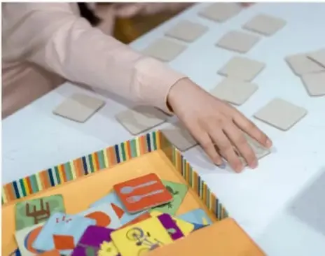 Fotografía de manos de un estudiante volteando fichas de un juego de memorama sobre una mesa; al lado hay la caja del juego con fichas de colores.