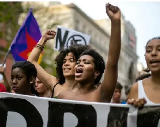 Fotografía de una manifestación de mujeres afrodescendientes con puños en alto, banderas y carteles que dicen NO, representando la lucha contra el racismo.