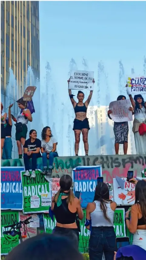 Fotografía de una manifestación pacífica con personas jóvenes sosteniendo carteles con mensajes como 'Escúchame, el nombre de tu homofobia/transfobia/lesbofobia es violencia' y 'Ternura radical', en un espacio público con una fuente al fondo.