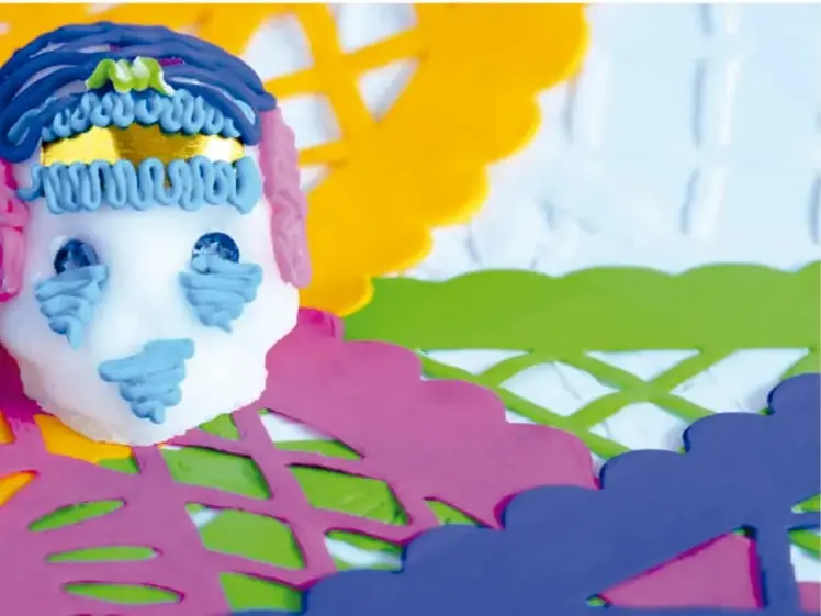 Calaverita de azúcar decorada en azul y dorado sobre papeles picado de colores (verde, morado, amarillo).