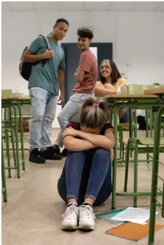 Estudiantes en un aula; una joven sentada en el suelo con la cabeza baja, mientras otros compañeros ríen al fondo.