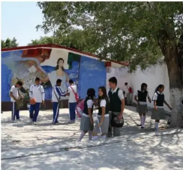 Fotografía de un grupo de estudiantes de secundaria caminando frente a un mural escolar al aire libre, con mochilas y uniformes. Representa la participación juvenil y la vida comunitaria en el espacio escolar.