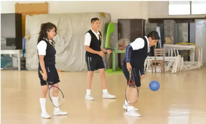 Tres estudiantes de uniforme con raquetas de tenis y una pelota azul en el patio o gimnasio de la escuela, ilustrando la actividad física lúdica.