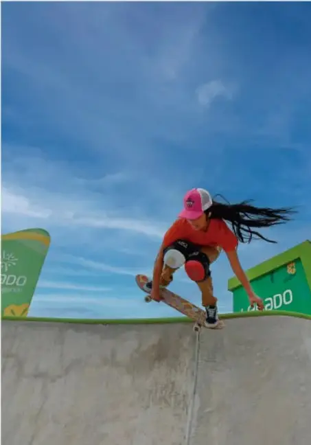 Fotografía de una joven con patineta, casco y rodilleras en un skatepark, bajo cielo azul, ilustrando la práctica de actividades recreativas.