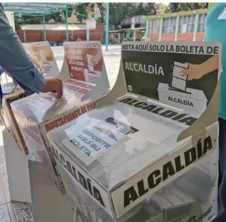 Módulo de una casilla electoral con carteles que dicen 'ALCALDÍA' y 'META AQUÍ SOLO LA BOLETA DE ALCALDÍA', con una persona depositando su voto.
