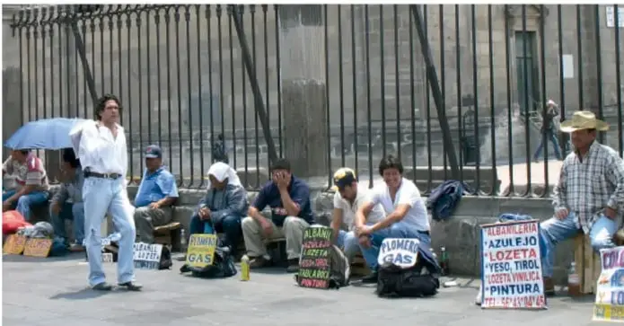 Fotografía de un grupo de trabajadores informales sentados en la banqueta frente a una reja metálica, con carteles que ofrecen servicios como albañilería, plomería, pintura y carga. Refleja la falta de oportunidades laborales formales.