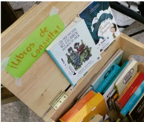Fotografía de una caja de madera con libros apilados y una etiqueta verde que dice "¡Libros de consulta!" en una biblioteca.