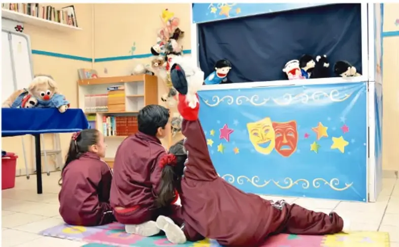 Fotografía de un aula de primaria: tres alumnos sentados en el piso frente a un teatrino de títeres con telón azul decorado con máscaras de comedia y tragedia y estrellas; títeres asomados por la apertura superior y un títere adicional sobre una mesa lateral.