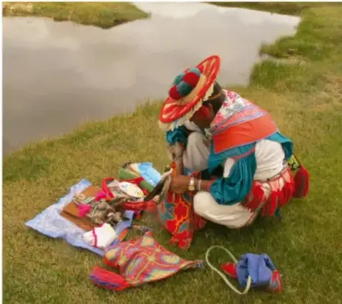 Fotografía de una persona indígena con atuendo tradicional multicolor arrodillada junto a un río, colocando ofrendas sobre una tela en el suelo, en un entorno natural de pastizal.