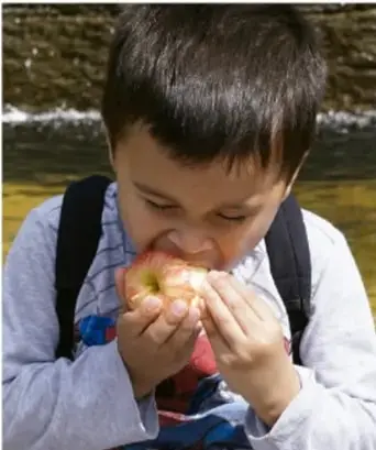 Niño comiendo una manzana con mochila, en un entorno al aire libre.