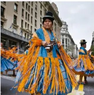 Fotografía de una mujer indígena con vestimenta tradicional de colores vivos durante una celebración en una calle de la ciudad.