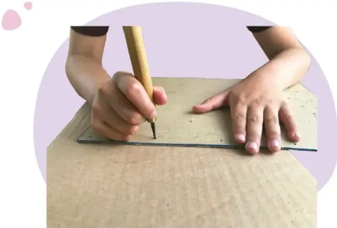Manos de un adulto perforando con un punzón un cartón apoyado sobre una regla, mientras la otra mano sujeta el material.