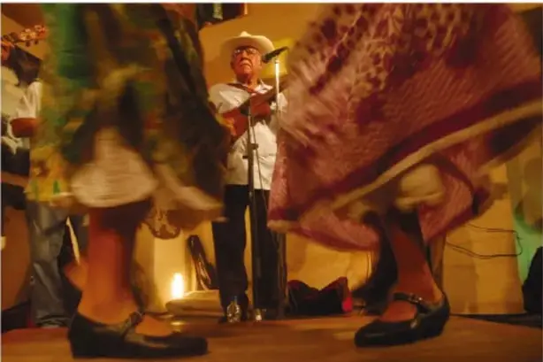 Artesano con sombrero de paja pintando una artesanía colorida en su taller, con máscaras y esculturas blancas de fondo; probablemente artesano de Veracruz o región jarocha.