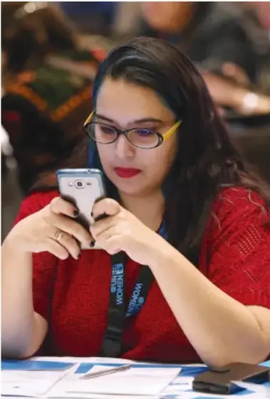 Joven con lentes y suéter rojo usando un teléfono inteligente, con expresión concentrada.