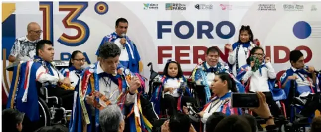 Fotografía de un grupo de personas, varias en sillas de ruedas, con sashes y distintivos multicolores, participando en un evento al aire libre con rótulo 'FORO' visible al fondo.