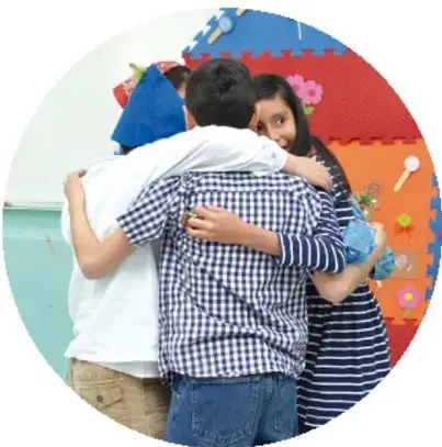 Tres personas (dos niños y una niña) abrazándose en un salón con piso de ecodes блоки de colores, transmitiendo convivencia y afecto.