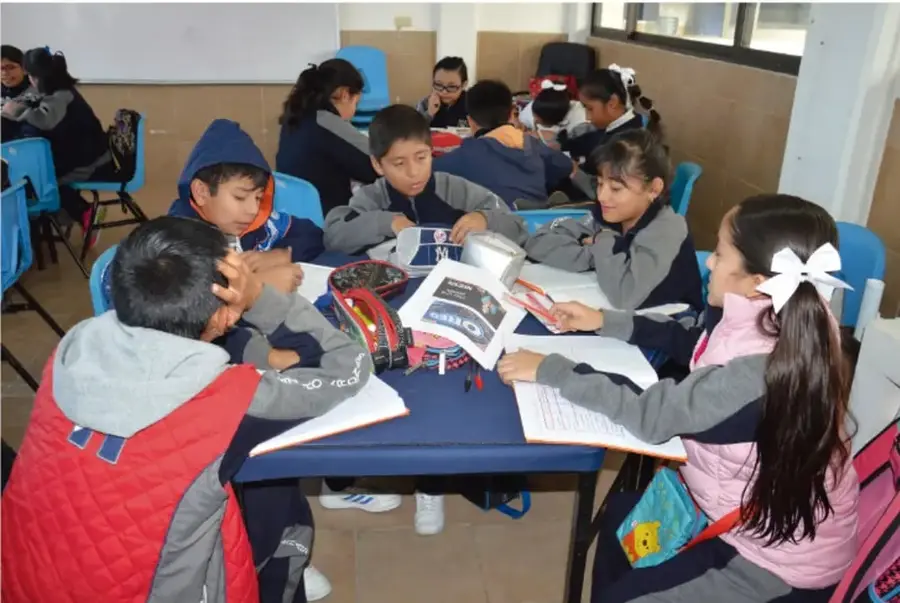 Fotografía de un grupo de estudiantes de primaria sentados alrededor de una mesa en el aula, revisando cuadernos y materiales; al fondo hay más compañeros trabajando en equipo.