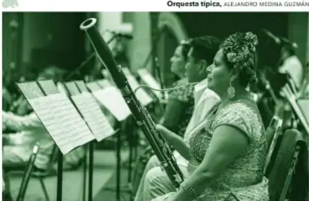 Fotografía en tono verde de una orquesta: al frente, una mujer con ropa típica, cabello trenzado con flores de tela, sostiene con la mano derecha un fagot; detrás se ven otros músicos con partituras en atriles.