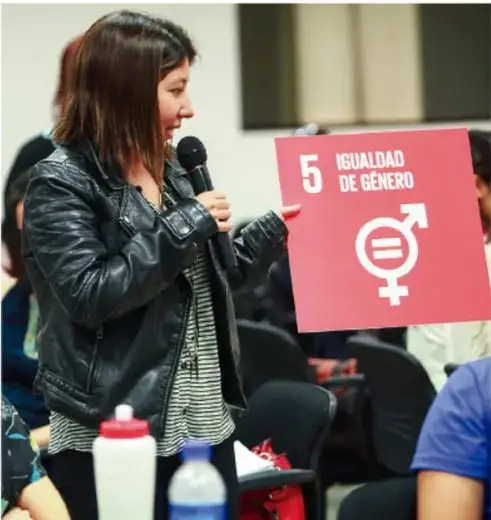 Mujer joven hablando con micrófono y sosteniendo un cartel con el número 5 y el símbolo de igualdad de género.