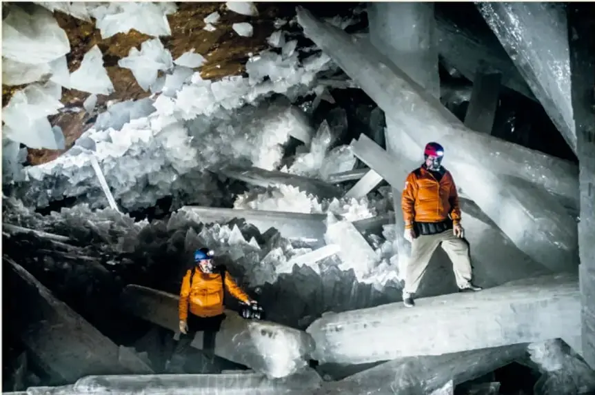 Fotografía de dos investigadores con trajes naranjas y cascos dentro de la Cueva de los Cristales, rodeados de enormes vigas de cristal blanco.