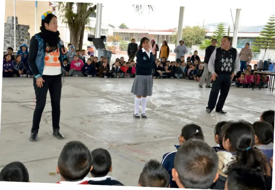 Fotografía de una representación escolar al aire libre: una maestra con micrófono, una alumna actuando y un adulto frente a un público de niños sentados en el patio.