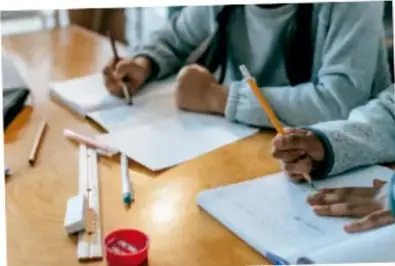Fotografía de dos estudiantes escribiendo con lápices en hojas sobre una mesa de madera.