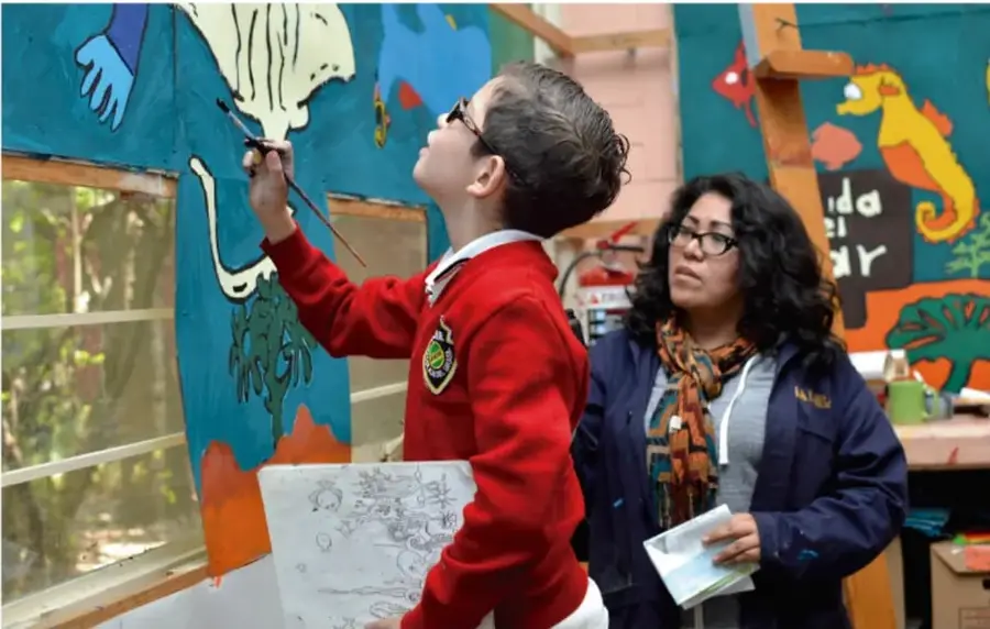 Fotografía de un niño con uniforme escolar rojo pintando un mural azul con un pincel, acompañado por una mujer adulta que sostiene un cuaderno; al fondo se observan dibujos de animales marinos y la palabra 'MAR'.