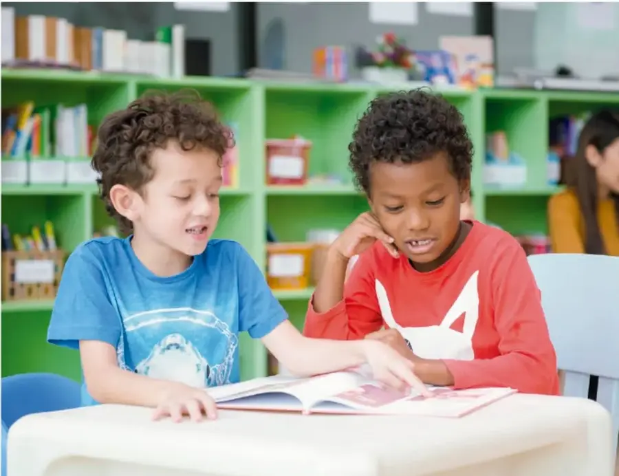 Fotografía de dos niños sentados en una mesa de la biblioteca escolar; uno de ellos señala un libro abierto mientras el otro observa y escucha. Al fondo se ven estanterías verdes con libros.