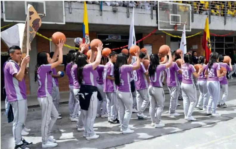 Fotografía de un grupo de estudiantes en uniforme deportivo morado, de espaldas, sosteniendo balones de básquetbol elevados sobre sus cabezas en un patio escolar con banderas.