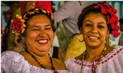 Fotografía de dos mujeres indígenas sonrientes con trajes tradicionales y flores en el cabello, ilustrando la diversidad cultural de México.