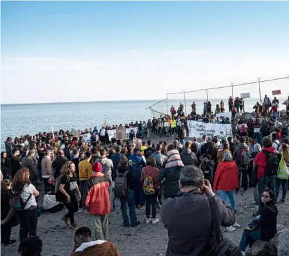 Fotografía de una multitud de personas reunidas en una playa junto a una valla metálica, con pancartas; al fondo el mar y un cielo despejado. Parece una manifestación o concentración en una frontera costera.
