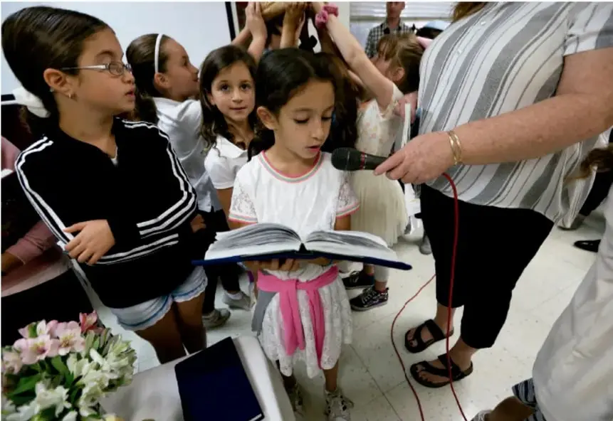 Fotografía de una niña de primaria leyendo en voz alta frente a un micrófono, acompañada por una maestra y compañeras de clase en un salón.