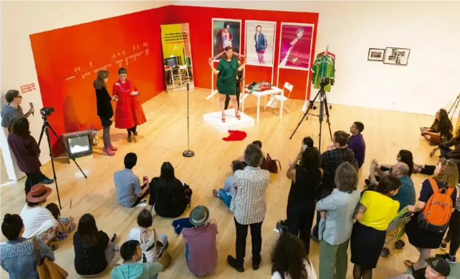 Fotografía de una acción performática en un espacio expositivo de paredes rojas: una persona actúa al centro, público sentado en el suelo observa y varias cámaras de video registran la acción.