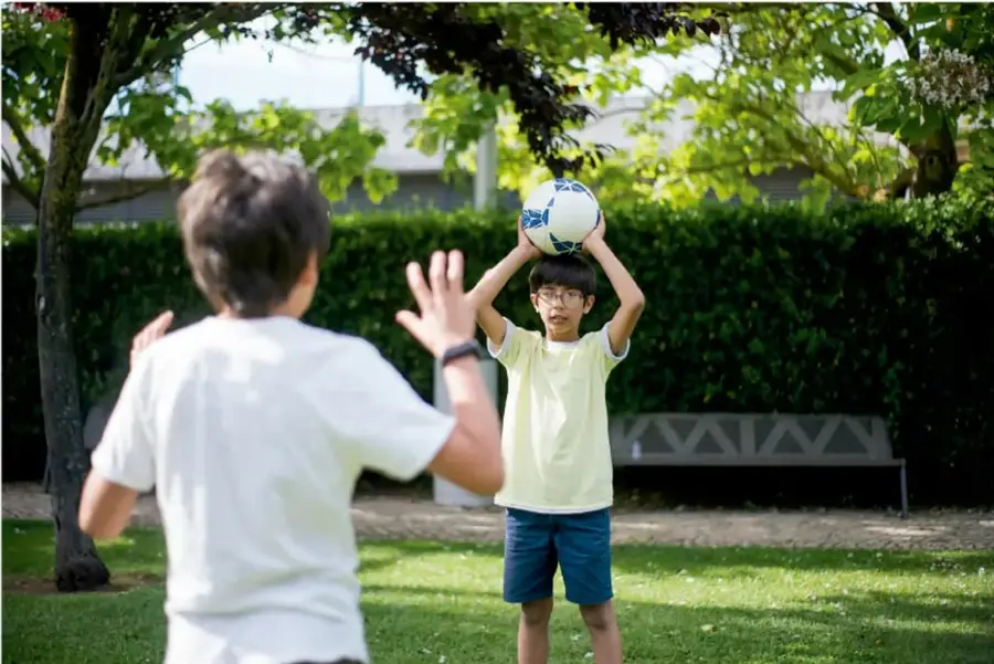 Fotografía de dos niños jugando fútbol en un parque. Uno sostiene el balón con las manos sobre la cabeza y el otro está de espaldas con los brazos abiertos.