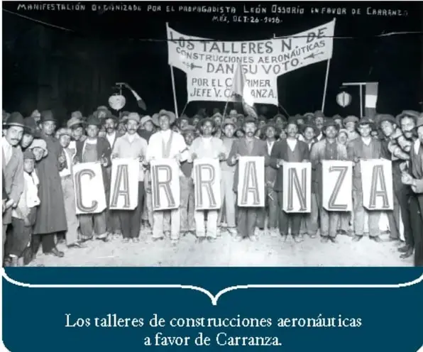 Fotografía en blanco y negro de una manifestación de 1916 a favor de Carranza; los asistentes sostienen letreros con la palabra CARRANZA y una manta de los talleres de construcciones aeronáuticas.