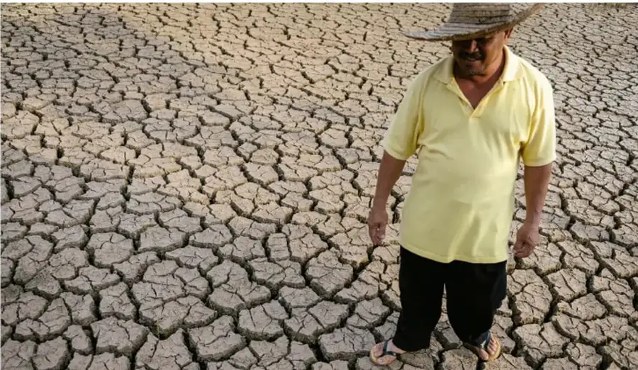 Fotografía de un agricultor con sombrero parado sobre suelo agrietado y seco, ilustrando los efectos de la sequía y el cambio climático.