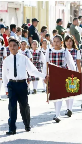 Estudiantes desfilando con uniforme escolar; una niña porta el estandarte de la escuela.