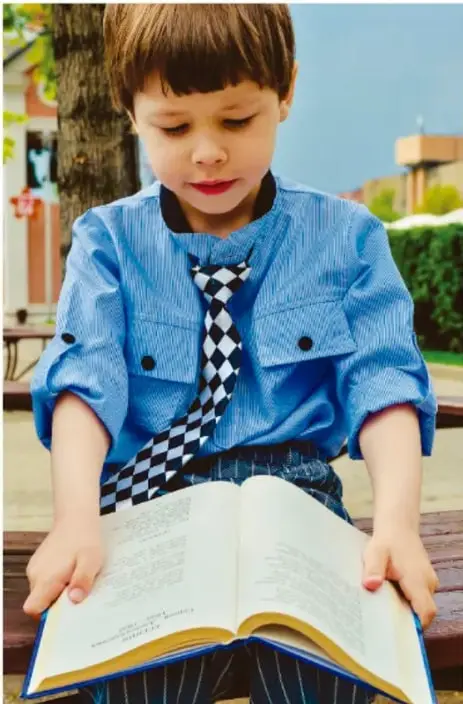 Niño con camisa azul y corbata de cuadros leyendo un libro abierto al aire libre.