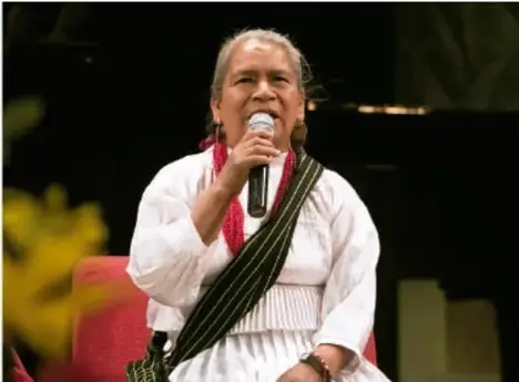 Mujer de cabello blanco con traje tradicional indígena y rebozo, sosteniendo un micrófono mientras habla en un escenario.