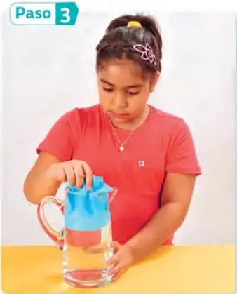 Niña con polera roja vierte agua de una jarra sobre un trozo de papel azul dentro de un recipiente de vidrio.
