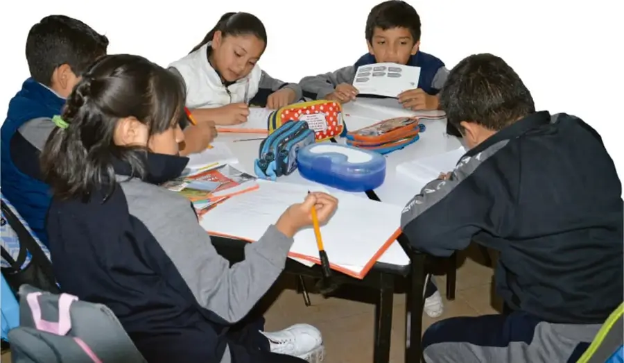Fotografía de cinco estudiantes de primaria sentados alrededor de una mesa blanca, trabajando en equipo con cuadernos, colores, una lonchera roja y una caja azul. Uno de ellos sostiene una hoja con recuadros.