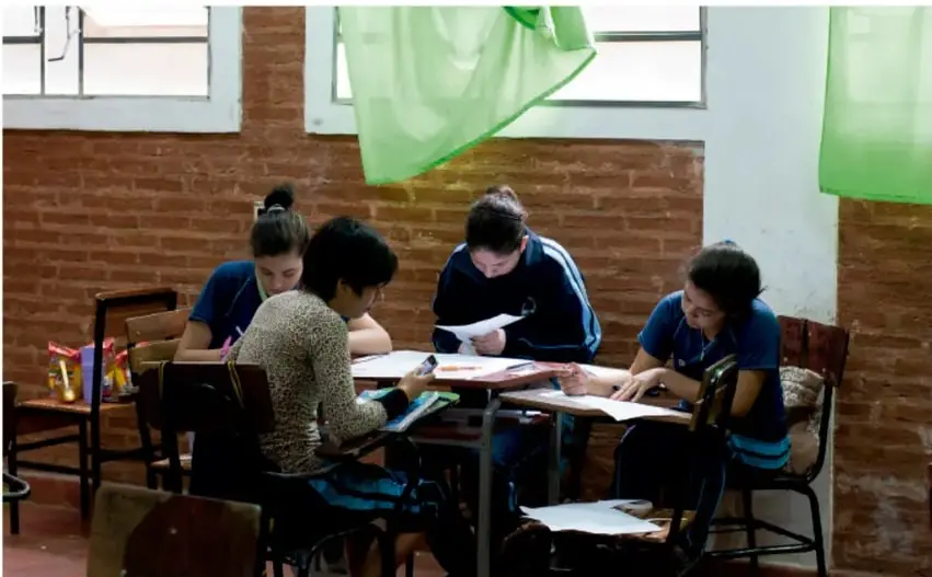 Fotografía de cuatro estudiantes de telesecundaria sentados alrededor de una mesa de trabajo en un aula de paredes de ladrillo, revisando hojas y fichas de papel: ilustran la elaboración colaborativa de los productos del proyecto.