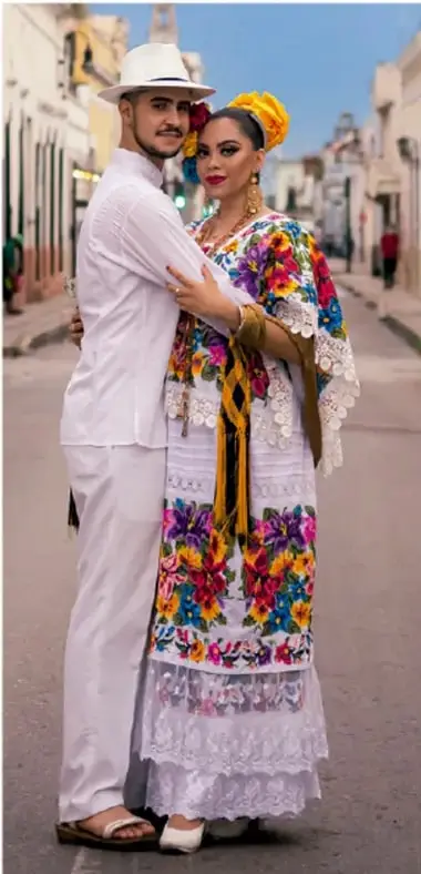 Fotografía de una pareja con traje tradicional mexicano: el hombre viste camisa y pantalón blancos con sombrero de paja; la mujer viste huipil blanco bordado con flores de colores y rebozo, en una calle colonial.