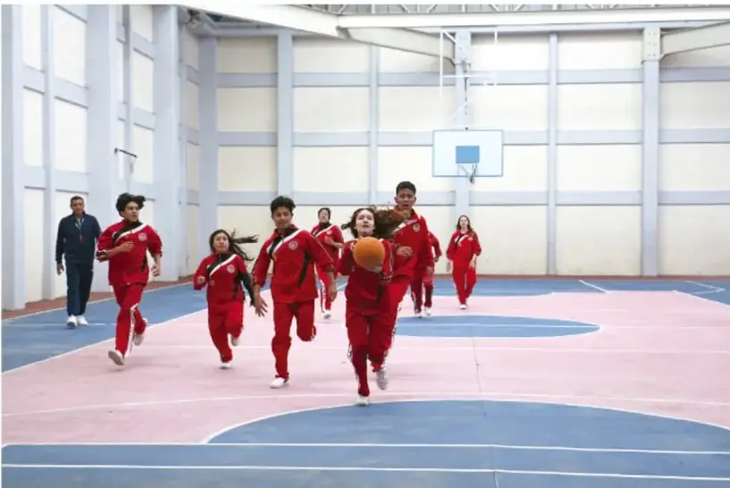 Grupo de estudiantes con uniforme rojo corriendo en un gimnasio cubierto durante una actividad física, con un profesor observando al fondo.