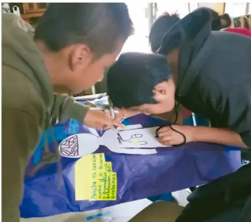 Fotografía de estudiantes trabajando con un mapa o boceto sobre un mantel azul, colaborando en equipo con notas adhesivas.