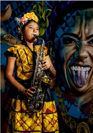 Fotografía de una joven adolescente tocando un saxofón, con vestimenta colorida de estampado tradicional (pañuelo en la cabeza con flores), frente a un muro con arte urbano que muestra un rostro pintado; ilustra la expresión a través del arte y la música.