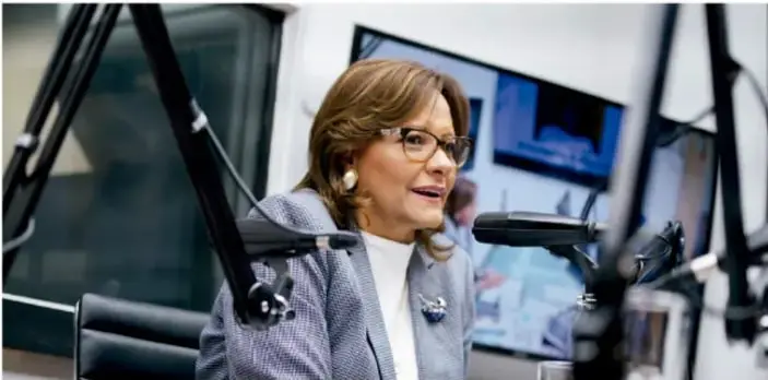 Una mujer con lentes y saco gris habla frente a un micrófono en un estudio de radio o televisión, con pantallas al fondo.