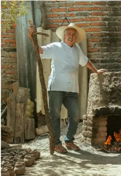 Fotografía de un artesano con sombrero, junto a un horno de ladrillo en funcionamiento, rodeado de piezas de cerámica recién horneadas.