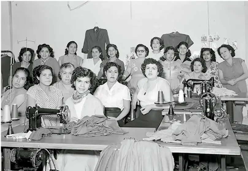 Fotografía en blanco y negro de un taller de costura con varias mujeres trabajando en máquinas de coser, algunas de pie y otras sentadas, con prendas de vestir sobre las mesas.